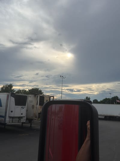 The image is a photograph taken from the perspective of someone in a red truck, looking out through the side mirror. The mirror reflects a portion of the truck's red exterior and a hand resting on the door frame.
The scene outside the truck appears to be a parking lot. Several large white trailers are parked in the lot, some with visible logos and markings. One trailer on the left has a blue logo. In the background, trees and streetlights are visible. Two American flags are also present in the distance, suggesting a location in the United States.
The sky is overcast with dramatic clouds, but a bright spot of sunlight is visible through a break in the clouds, creating a dramatic effect. The ground is asphalt. The overall impression is that of a truck stop or industrial area, possibly in Murfreesboro, USA, given the context provided. The time of day appears to be late afternoon or early evening due to the lighting conditions.
