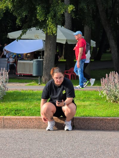 In Veliky Novgorod, Russia, a woman with glasses and reddish-brown hair tied up is squatting on a sidewalk in a park. She is wearing a black t-shirt and black shorts. She is holding a black smartphone in her right hand and adjusting the laces of her white athletic shoe with her left hand. In the background, a man in a red t-shirt and blue jeans is walking, and behind him, there is a white tent with the "CARTE D'OR" logo. The park has green grass, trees, and a granite border along the sidewalk.