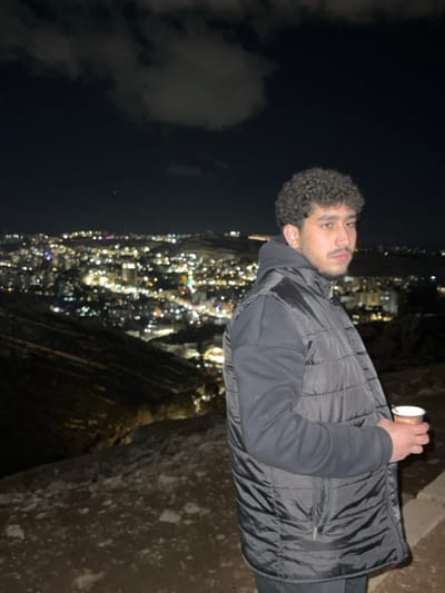 A young adult male is depicted at night, standing on a rocky incline overlooking the city of Dummar Municipality, Syria. The male, positioned slightly right of center, faces left with his head turned towards the viewer. He has dark, curly hair and a beard, wearing a dark hooded sweatshirt under a dark, quilted vest. His right hand holds a white paper cup with brown/orange markings. The background features a sprawling cityscape illuminated by numerous blurred lights, indicating a significant distance and nighttime conditions. Dark, undulating terrain is visible between the male and the city lights. The sky above is dark with faint cloud formations. The immediate foreground consists of rugged, dark earth and rocks.