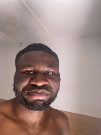 The image displays a close-up portrait of an adult male in an indoor environment located in Udu, Nigeria. The individual, appearing to be of African descent, has a dark skin tone, short dark hair, and a full dark beard and mustache. His dark eyes are directed forward, and his lips are subtly downturned, indicating a neutral facial expression. He is depicted from the shoulders up, appearing shirtless. The background features a predominantly white wall and ceiling. The ceiling shows faint brown discoloration, particularly on the upper left, along with visible linear seams. A small, dark, circular object is affixed to the upper left section of the wall. A horizontal strip of light brown or beige material is visible on the lower right wall. The subject is looking directly at the camera, suggesting a posed photograph or selfie.