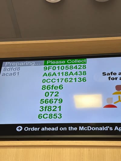 The image shows a digital screen at a McDonald's location, possibly in or near Pity Me, United Kingdom, although the exact location cannot be determined from the image alone. The screen displays order preparation and collection information. On the left, the text "Preparing..." appears, followed by the alphanumeric code "8dfd8 aca61." In the center, under the heading "Please Collect," a list of alphanumeric order codes (9F01058428, A6A118A438, 0CC1762136, 86fe6, 072, 56679, 3f821, 6C853) is displayed. To the right of the order codes, there's a visual element suggesting a safe and the wording "Safe a for a". At the bottom, an arrow icon precedes the text "Order ahead on the McDonald's Ap." The screen is framed by a wooden surface.