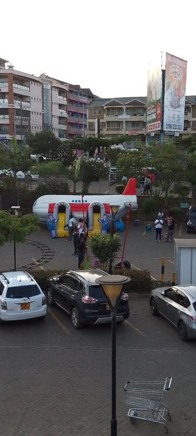 The image depicts an outdoor urban environment located in Ngong ward, Kenya. A paved central area features an inflatable bounce house designed as a white and red passenger airplane, equipped with two yellow slides and two inflatable figures resembling airline staff at the base of the slides. Approximately eight to ten individuals, comprising both adults and children, are present around and within the inflatable structure, engaged in recreational activity. Sections of a miniature train track are visible on the paved ground to the left of the inflatable.
In the foreground, a paved parking lot contains three parked vehicles: a white Toyota SUV/wagon (license plate KCH 433V), a dark grey SUV (partial license plate K*** 84), and a silver sedan. A black street lamp post and a metal shopping cart are positioned in the lower foreground. The background is dominated by multi-story buildings, exhibiting various architectural styles and colors, indicative of residential or commercial use. A large, illuminated billboard on the right side of the background displays advertisements, including the text "MUTHOKINJU," contact numbers "0722 99 30 30" and "0790 022 022," and "50x100 PLOTS IN NGONG," providing specific location context. Trees and shrubs are interspersed throughout the scene, separating structures and parking areas. The sky is clear, indicating daylight conditions.