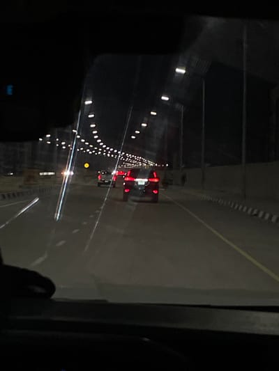 The image depicts a nighttime scene from the interior of a moving vehicle on a multi-lane roadway in Alaguntan, Nigeria. The perspective is through the front windshield, exhibiting reflections from internal dashboard elements and external streetlights. Ahead, three vehicles are visible; the closest is a dark sport utility vehicle (SUV) with illuminated red tail lights, followed by a lighter-colored pickup truck, and another vehicle further in the distance. The road is asphalt, marked with white dashed lane lines and a solid yellow line on the right shoulder. Overhead, numerous streetlights arranged in an arc illuminate the thoroughfare. Black and white striped concrete barriers delineate the left and right sides of the road. On the far right, alongside the barrier and a dark wall, a lone, indistinct dark figure, possibly a person, is visible. The environment is dark, suggesting a clear night with artificial illumination as the primary light source.