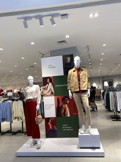 This image captures the interior of a brightly lit retail clothing store in Bekasi Regency, Indonesia, with the camera oriented in a portrait aspect but rotated 90 degrees clockwise. The scene features two mannequins positioned on elevated white platforms.

In the foreground, a male mannequin on the left wears a beige jacket over a red patterned shirt and light-colored trousers, paired with white sneakers. To its right, a female mannequin is dressed in a light-colored, long-sleeved blouse, maroon culottes, and light-colored open-toed shoes, holding a small beige handbag.

Behind the mannequins, a large vertical display board is visible, divided into multiple panels featuring images of people and text. Visible text elements include: "art is theraphy.", "where words fail, music speaks.", "Everyone Exe", and "I don't do fashion, I am fashion."

Further into the background, racks of clothing, both hanging and folded, are arranged, showcasing various garments. The store's ceiling is equipped with numerous recessed circular lights and several white track lighting fixtures, illuminating the space evenly. The overall setting is that of a modern and organized fashion retail environment.
