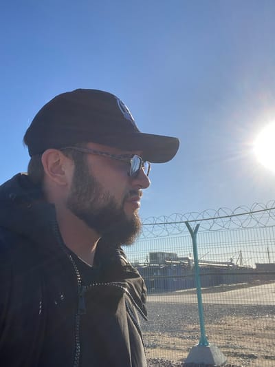 The image shows a man wearing a black baseball cap and sunglasses, looking to his right. He has a beard and is wearing a black jacket. Behind him, a barbed wire fence separates him from a background that appears to be an industrial area with shipping containers and buildings. The sky is bright blue with the sun shining, creating a lens flare on the right side of the image. The location is stated to be the city of Koskolovo, Russia.