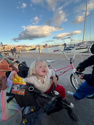 The image presents a detailed view of a harborside promenade in Aegina, Greece, under a partly cloudy sky during late afternoon or early evening.

In the foreground, a young child with blonde hair, dressed in a light-colored long-sleeved top and red trousers, is seated in a black stroller marked "ROYAL." The child's mouth is slightly open, and they hold a yellow "Haribo Goldbären" snack bag in their right hand. A brown, fox-like stuffed animal rests to the child's left within the stroller, which also contains a rainbow-patterned blanket and a dark bag adorned with yellow star-shaped accessories. To the immediate right, the lower torso and right arm of an individual, wearing a dark top, blue trousers, and light-colored sneakers, are visible. This individual's hands are positioned near the handlebars and front wheel of a pink "HERO" brand BMX-style bicycle with white components. The subjects are positioned on a grey paved surface consistent with a pedestrian walkway.

The midground features the Aegina harbor, where numerous sailboats and at least one white motor yacht are moored. Their masts are prominent against the sky. Along the waterfront, a continuous line of multi-story buildings with traditional Greek architecture and varied roof colors, including terracotta, is visible. A distinctive building with a red dome is situated on the far right of the waterfront structures. To the left of the main subjects, another individual is seen walking alongside a light-colored bicycle equipped with a rear basket. Distant mountains are faintly discernible behind the town. The sky is clear blue with scattered white cumulus clouds.