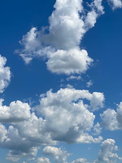 The media file presents a clear, bright blue sky filled with numerous cumulus clouds. The clouds vary in size and density, appearing predominantly white with some gray undertones, indicative of direct sunlight from above and shadow play within their forms. They are clustered on the left and right sides of the frame, with a central strip of deep blue sky. There are no people, objects, or specific activities depicted. The scene captures an outdoor setting on what appears to be a sunny day, likely mid-day given the intense blue of the sky and the bright illumination of the clouds. No location cues are visible within the image itself, but the context provided is Zaķumuiža, Latvia. There is no visible text.