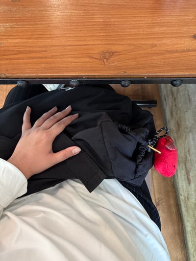 This is a close-up, top-down, vertically oriented shot of a person's hand resting on a pile of black and white clothing, situated next to a wooden surface. The hand, with clean, well-maintained fingernails, is gently placed on a gathered black fabric, possibly a jacket or trousers. To the left of the clothing is a vertical wooden surface with a visible grain and two dark, round rivets on a black metallic strip running along its edge. Above the clothing, a small, bright red plush toy with a checkered ear is attached to a drawstring with a black and white cord. The red toy is positioned on top of the black clothing. A thin golden rod or skewer is inserted through the drawstring of the black garment, near the toy. A hint of the black and white pinstriped pattern of another garment is visible beneath the black fabric, and a white sleeve is also present. The scene appears to be indoors, possibly in a residential setting, given the casual arrangement of clothing and the presence of a toy. The lighting suggests it could be daytime, but no specific time is discernible. No text is visible in the image. The location context of Chiang Mai City Municipality, Thailand, does not provide specific visual cues within the frame.