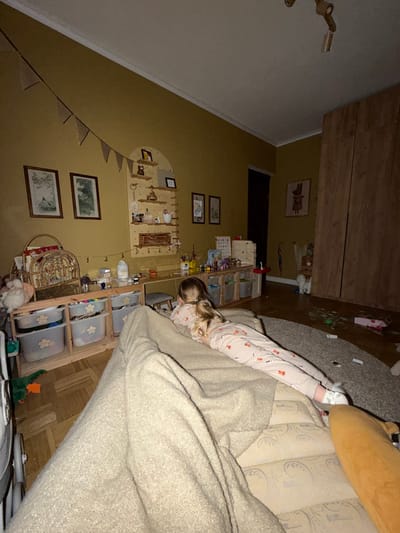 This image depicts an indoor domestic scene in Искра Снт (чашниково), Russia, characterized by subdued artificial lighting. The room's walls are painted a uniform mustard yellow, contrasting with a white ceiling and crown molding. The floor is covered with light-colored wood planks in a herringbone pattern on the left and a light grey textured rug on the right.
A young child, appearing female with long blonde hair tied in a ponytail, is the sole visible individual. She is lying prone on a light-beige upholstered sofa or bed in the foreground, facing away from the camera, and looking towards the far wall. She is dressed in light-colored pajamas adorned with a repeating pattern, possibly foxes, and white socks or slippers. A beige textured blanket is draped over the sofa, partially covering her.
Along the main wall, an arched wooden shelving unit holds various decorative items and small bottles. Below it, a long, low wooden desk or table extends, featuring numerous art supplies, toys, and miscellaneous objects on its surface. Underneath the desk, multiple light-grey plastic storage bins with flower-shaped cutouts are organized within wooden cubbies. A small grey-cushioned stool is tucked beneath the desk. To the left of this setup, two framed botanical prints are hung on the wall. A string of small, triangular pennants hangs diagonally from the upper left corner of the room towards the arched shelving.
To the right, a dark vertical opening, likely a doorway, is visible. Adjacent to it stands a tall, light-colored wooden wardrobe with two panel doors. Between the doorway and the wardrobe, a framed print of a stylized rabbit or animal is displayed on the wall. On the floor, near the wardrobe and on the rug, various items are scattered, including a white stuffed animal, a small red umbrella, and several indistinct objects, with what appears to be puzzle pieces on the rug. A green toy, possibly a duck or dinosaur, rests on the wooden floor in the midground. A brown/orange stuffed animal or cushion is partially visible on the far right of the sofa in the immediate foreground.