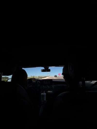 The image depicts the interior of a moving vehicle, viewed from a rear passenger perspective, looking forward through the windshield. The scene is located on a highway in El Paraíso, Mexico.

**People:**
- A person with dark hair is in the driver's seat on the left, with their hands on the steering wheel, indicating active driving. Their face is obscured by shadow.
- A second person, wearing a light-colored cap, occupies the front passenger seat on the right. Their back and head are visible, also heavily shadowed.

**Vehicle Interior:**
- The interior of the car is predominantly in deep shadow, with only the silhouettes of the occupants and dashboard visible.
- The dashboard is modern, featuring an infotainment screen, circular air vents, and various controls.
- A light-colored travel mug or cup is secured in a cup holder on the center console. A dark object, possibly a strap or umbrella, hangs adjacent to the cup holder.
- A rearview mirror is visible at the top center of the windshield opening.

**Exterior Environment:**
- The vehicle is traveling on a multi-lane highway under a clear, bright blue sky.
- A concrete or asphalt road surface is visible with white lane markings.
- A white and red striped concrete barrier or guardrail runs along the right side of the road.
- Green vegetation, consistent with bushes and trees, lines both sides of the highway.
- In the distance ahead, a dark-colored vehicle is visible in the same lane, and another, less distinct vehicle, is further ahead.
- A prominent pink billboard with white text is visible on the right side of the road in the distance.
- Utility poles or streetlights are visible alongside the highway.