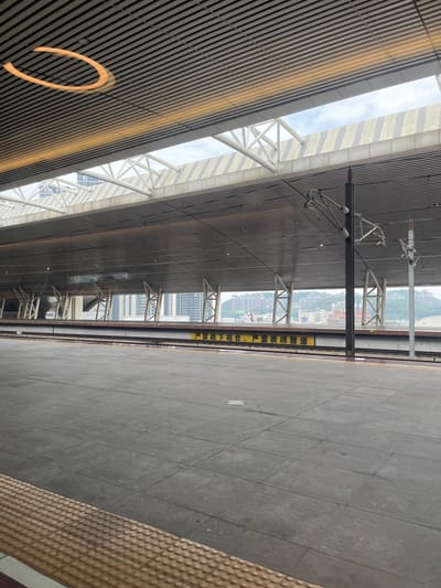 This image depicts the interior of a modern train station platform in Dongguan, China. The scene is dominated by the station's architecture, featuring a large, sloped roof made of horizontal wooden slats and supported by white metal trusses. A bright sky is visible through large open sections of the roof, suggesting daytime with possibly overcast weather. The platform itself is paved with grey tiles, with a textured yellow tactile paving strip along the edge.
Several objects are visible. Overhead, a complex system of catenary wires and supporting poles, typical of an electrified railway, stretches across the platform. A vertical sign with yellow text on a dark background is attached to one of these poles. In the distance, through the open roof structure, a cityscape with numerous buildings and a hilly landscape can be seen. The lighting appears to be natural daylight, casting subtle shadows.
The image is taken from a slightly elevated perspective, looking down the length of the platform. The overall impression is one of efficient, modern public infrastructure. No people are visible in the frame, and the station appears quiet. The prominent yellow text on the sign reads "严禁烟火" and "下车请注意安全", which translates to "No smoking" and "Please pay attention to safety when alighting".