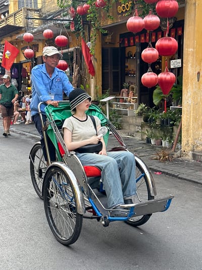 The image depicts a street scene in Hội An Ward, Vietnam, characterized by a weathered yellow building and a grey asphalt street. Red lanterns are strung across the street, adding a festive touch.

The central focus is a cyclo, a three-wheeled vehicle propelled by a rider. A man in a blue shirt and a white cap is operating the cyclo, facing forward with a neutral expression. Seated behind him, in the passenger compartment, is a young person wearing a light-colored t-shirt and a black and white striped beanie. This passenger appears to be resting or sleeping, with their head tilted back and eyes closed. A small white fan is clipped to the side of the cyclo, suggesting a warm day. The passenger is wearing blue jeans.

In the background, several shops are visible, adorned with flags featuring a yellow star on a red background, likely Vietnamese flags. One shop has a sign that reads "COFFEE." There are also shelves displaying various items, possibly souvenirs or goods for sale. The overall impression is a snapshot of daily life in a bustling tourist area, with the cyclo serving as a mode of transportation and a symbol of the local culture. The lighting suggests it is daytime.
