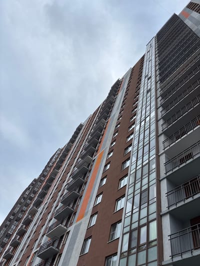 A low-angle shot depicts a tall, multi-story residential building in Kudrovo, Russia. The building is a mix of brown and grey facades with windows and balconies, reaching toward a cloudy, overcast sky. Balconies appear on both sides of the building.
