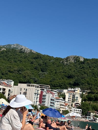 The image depicts a vibrant, sunny beach scene in Rafailovići, Montenegro. A clear, expansive blue sky dominates the upper portion of the frame. Below the sky, a large, densely forested mountain range with visible rocky outcrops stretches across the background.
Mid-ground, between the mountain and the beach, several multi-story buildings, likely hotels or apartments, are constructed on the hillside. These buildings feature light-colored facades, multiple balconies, and some red and white accents.
The lower half of the image focuses on a crowded beach. In the immediate foreground, facing away from the viewer and slightly to the left, is a woman wearing a large white wide-brimmed hat and a white sheer shirt. Her right hand is raised to her face, partially obscuring it. She appears to be seated on the sand or a low beach chair.
Behind her, the beach is populated with numerous individuals sunbathing, sitting, or wading in the water. Various beach umbrellas of different colors, including blue, white, and multi-striped, are scattered across the sand, providing shade. A blue umbrella is prominently featured to the left of the main subject, and a white one is further to the right. To the right of the woman in the white hat, a bald man is visible, reclining on the beach. More people are indistinctly visible further down the beach and in the shallow turquoise water. The overall scene suggests a busy coastal resort environment during fair weather.
