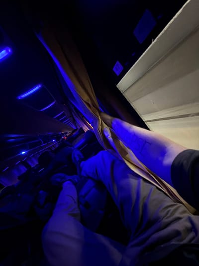 The image appears to be taken inside a dimly lit bus or train, with blue ambient lighting illuminating the interior. The perspective is from a passenger's viewpoint, looking down the aisle of the vehicle. Several seats are visible, some occupied and some empty, with yellow curtains partially drawn. In the foreground, a person's legs are visible, wearing light-colored pants and sneakers. The person's right arm is also in view, with the word "COLLAPSE" written on it in dark ink. The environment suggests a journey, possibly at night, given the low light conditions and the presence of overhead lights. There is no visible indication that this scene is specifically located in Bilaspur, India.