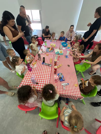 This media file captures a gathering of adults and children around a table covered with a red and white checkered tablecloth. The subjects are primarily young children seated at small, brightly colored plastic chairs, interacting with snacks and drinks laid out on the table. Several adults are also present, observing and possibly assisting the children.
The scene appears to be an indoor setting, likely a community center or a home, given the plain gray floor and the presence of windows in the background. The overall atmosphere seems relaxed and informal, suggesting a party or a shared meal. The children are engaged with their food, some are eating, while others are looking at the items on the table.
Notable details include various food items and beverages spread across the table, such as waffles, candies, and what appear to be juice boxes or water bottles. Some of the children are holding these items, indicating they are actively participating in the event. The clothing of the subjects is casual, consistent with a daytime gathering.
There is no visible text in the image that provides specific information about the event, time of day, or weather. However, the bright lighting suggests it is daytime. The emotions conveyed are generally positive, with children appearing engaged and adults seemingly content. The context provided indicates the location is Kiryat Bialik, Israel.