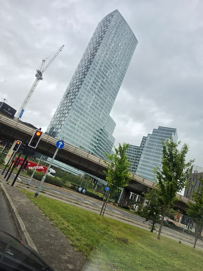 The image captures an overcast urban scene in Manchester, United Kingdom, specifically depicting the area around **Deansgate Square** and **Spinningfields**. The perspective is from a street level, looking upwards at modern architecture.
A dominant feature is a tall, curved, glass-clad skyscraper, identified as **One Spinningfields**, situated prominently on the left-center, its reflective surface mirroring the grey sky. To its right, a substantial elevated concrete flyover or motorway (likely part of the Mancunian Way) traverses horizontally across the middle ground. Beyond this elevated structure, several other contemporary glass-fronted buildings with a stepped design, characteristic of the **Deansgate Square** development, are visible under the overcast sky.
On the far left, a tall white lattice construction crane extends upwards, signifying ongoing development. Below the crane, a dark building with "VITA STUDENT" signage is partially visible. In the immediate foreground on the left, three sets of traffic lights display red (or orange-red) signals, next to a blue circular road sign indicating a straight-ahead direction.
Beneath the elevated road, at ground level, a multi-lane asphalt road is visible. A red vehicle, possibly a truck or bus, bearing "T.K.maxx" and "60%" advertising, is partially seen on the road, alongside a white van. Two indistinct human figures are walking on the road or pavement beneath the flyover. The foreground includes a grey block-paved sidewalk, strips of green grass, and several young, leafy green trees. The bottom-left corner shows the dark roof and interior elements of the vehicle from which the photograph was taken.