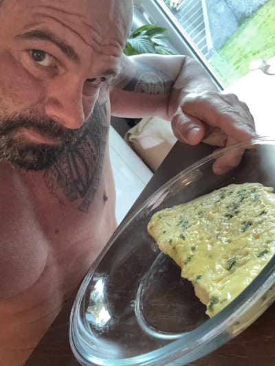 A shirtless man with a beard and tattoos on his chest and arm holds a clear glass dish containing a partially eaten omelet with visible green herbs, likely chives. The background is blurred, but a window and a plant are visible. The image appears to be taken indoors, possibly in a domestic setting. There is no indication of the city of Marche-en-Famenne, Belgium, in the image.