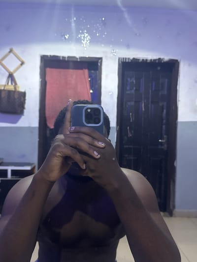 A mirror selfie captures a person holding a smartphone in an indoor setting. The subject, with dark skin, is visible from the mid-torso up on the right side of the frame, holding a blue/gray smartphone, which appears to be an iPhone model, featuring multiple camera lenses. The person's fingernails are painted a light pink hue with small black line accents. They are wearing a dark top or garment.
The background, which is the reflection of the room, features a light-colored wall, possibly white or very pale blue/gray, showing some texture and subtle variations in tone. On the left side of the reflection, numerous small, bright, circular bokeh lights are scattered, suggesting reflections from ambient light sources or decorative string lights.
Two dark, rectangular frames, likely made of wood and resembling door or window frames, are prominent in the background. The upper frame reveals a dark, indistinct area behind it, while the lower frame shows a reddish object, possibly a piece of fabric or furniture. Below the lower frame, towards the bottom left, a wooden, diamond-shaped decorative item or stand is partially visible, next to a brown patterned bag.
The overall lighting appears dim, suggesting an indoor environment, possibly in the evening or night. No discernible text is visible within the image. The scene is set indoors in Gbongan, Nigeria.