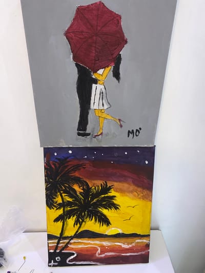 The image shows two acrylic paintings on canvas, positioned side-by-side on a white surface.
The left painting depicts a figure, presumably a woman, lying on a gray ground with a maroon umbrella obscuring much of her body. She is wearing a white and black striped skirt with a yellow top, and her shoes are a similar shade of maroon. The signature "mo." is visible near the top edge.
The right painting features a tropical beach scene at sunset or sunrise. There are silhouettes of palm trees in the foreground against a yellow and orange sky. The upper portion of the painting is dark blue with white spots. The signature or initials "zay. 2020." are located at the bottom left and a "+" symbol at the bottom right.
The location of Ibadan, Nigeria is not directly depicted in the image, however, the tropical beach scene on the right painting hints at a tropical or subtropical climate, and it is possible that the artist of the piece associates the artwork with Ibadan, Nigeria.