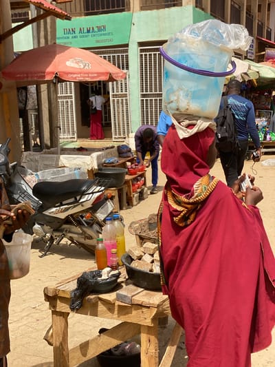 This sunlit image captures a bustling street market scene in Kafanchan B, Nigeria, during daytime. The scene is filled with people, market stalls, and various goods, indicating an active commercial environment.
In the midground, a person wearing a prominent red robe with a patterned trim on the sleeve is seen from behind, holding a small white object, likely a phone. To their left, another individual in a blue shirt holds a black object, possibly another phone. Further back, a person in a blue long-sleeved top is engaged near a market stall, and another individual in a purple top is bending over, seemingly inspecting items. In the background, a person in a red outfit stands near a building entrance.
Notable objects include a large, translucent light blue plastic bucket, partially covered by a clear plastic bag, which appears to be carried on someone's head. A black and white motorcycle is parked with various items on and around it, including several small plastic bottles containing colorful liquids (possibly beverages or oils) displayed on a wooden stand. A large red market umbrella, open for shade, stands prominently. Makeshift wooden structures and crates are used as market stalls or display areas.
The bright, direct sunlight suggests it is mid-day with clear weather. A light green building forms part of the backdrop, featuring a white grilled window. Visible text on the green building sign reads "DAN ALMAJIRI PROVISION AND GOOD ENTERPRISES" along with two phone numbers, "08032337243" and "7500579". The red umbrella clearly advertises "Original Tiger Blade Sold Here." The word "SOKOTO" is also visible on what appears to be a motorcycle's license plate. The overall activity suggests everyday commerce and social interaction within the market.