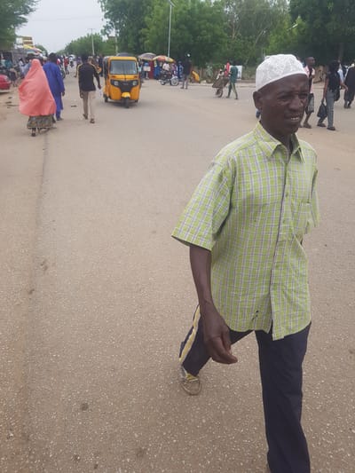 An elderly man wearing a white kufi cap and a plaid, short-sleeved green shirt stands on a paved road in Dabawa, Nigeria. He wears dark trousers with a yellow stripe near the hem and beige shoes. Other people walk in the background. A yellow tricycle taxi is parked on the side of the road. Trees and buildings are visible in the background. The road appears to be a main thoroughfare.