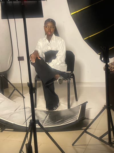 The image depicts a young Black woman posing in a studio setting in South Ropp, Nigeria. She is seated, leaning back on a chair, with her legs extended and crossed at the ankles. The woman is wearing a white collared shirt and black wide-leg trousers. She has her hair pulled back and is adorned with jewelry, including a necklace and earrings. Her nails are painted a light color.
The scene is illuminated by studio lighting equipment, including large softboxes, one of which is positioned to the left of the frame and another partially visible to the right. A black backdrop or large object is situated behind her, creating a stark contrast with the white wall behind. The presence of lighting equipment suggests a photoshoot is in progress. The overall mood appears to be calm and posed, with the woman looking directly at the camera. There are no visible text elements in the image.