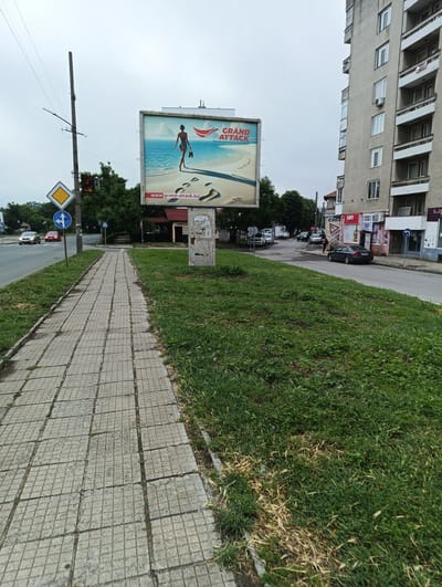 The image depicts a street scene in Karnobat, Bulgaria, on what appears to be an overcast day. A wide, grey tiled sidewalk extends from the lower left towards the mid-ground, flanked on its right by a strip of green grass. Beyond the grass and sidewalk, an asphalt road is visible, with several cars, both parked and moving.

A large billboard stands prominently to the right of the sidewalk. Its advertisement shows a woman in a blue swimsuit walking on a sandy beach towards the ocean, carrying sandals. Text on the billboard reads "GRAND ATTACK" and "www.grand-attack.bg," with footprints clearly visible in the sand.

To the left of the billboard, a tall utility pole supports a streetlight and various traffic signs. These include a yellow diamond sign indicating a priority road, and a blue circular sign with a left-turn arrow. In the background, a multi-story residential building with numerous windows and balconies rises on the right side of the street. Its ground floor features several commercial establishments with visible signs reading "CME," "КОМ," "EXIT," and "ХОРИЗОНТ" (Horizon).

Further down the street, more parked cars are visible, and a person is seen near a dark sedan. The scene suggests a typical urban environment with daily activity.