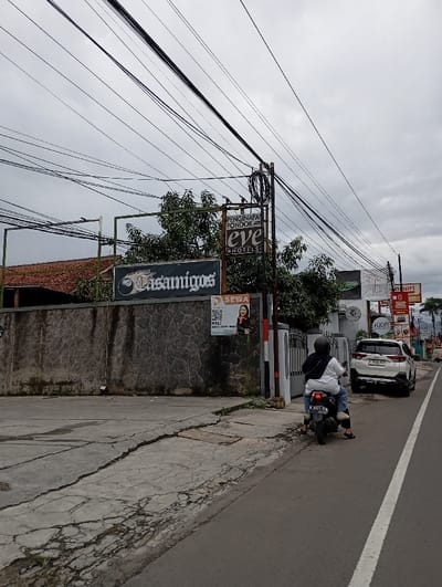 The image depicts a street scene in Katapang, Indonesia. The location is confirmed by the coordinates provided: -6.994 latitude, 107.555 longitude. The street is paved with asphalt and has a white lane divider. There are several utility poles with numerous power lines overhead. The street is lined with a gray concrete curb. A person is riding a motorcycle in the street. The person is wearing a gray jacket and a helmet. The motorcycle is black and white. Behind the motorcycle, there is a white car parked near a gray concrete wall. The wall has various signs and advertisements, including a sign for "Eve Hotels" and a sign for a business called "Casamigos." On the wall there is also a sign for "Sewa" with a woman's photo. The wall appears to be part of a building. Behind the wall is a section of greenery. The sky is overcast and gray. The overall impression is one of a typical street scene in an urban area.