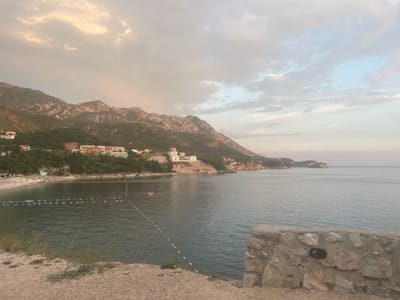 The image depicts a serene coastal scene at Kamenovo, Montenegro, likely during the late afternoon or early evening. The sky is partly cloudy with soft, diffused light, suggesting either an approaching sunset or a generally overcast day. The sea is calm and stretches out to the horizon, meeting a pale sky.
In the foreground, a low, rough stone wall lines the edge of a gravelly embankment, with some sparse grass growing near it. Beyond the wall, a beach is visible, lined with numerous white lounge chairs and umbrellas, indicating it's a tourist or resort area. The water near the shore is shallow and marked with buoys and ropes, delineating swimming areas. There are a few small objects in the water, possibly buoys or inflatable toys. A couple of small boats or watercraft are also visible.
Further back, nestled against the foothills of a mountain range, are several buildings. These appear to be hotels or residential complexes, with white and earth-toned facades. The mountains themselves are rugged and covered in green vegetation on their lower slopes, becoming more rocky and bare towards their peaks. The overall mood is peaceful and tranquil, with a sense of natural beauty and leisure. No visible text is present in the image.