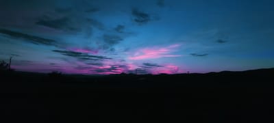 A wide-angle, low-light landscape photograph captures a dramatic twilight sky over Momin Prohod, Bulgaria. The sky is dominated by vibrant hues of deep blue, transitioning to streaks of electric pink and purple, indicating the lingering light of sunset or the approaching dawn. Wispy, dark clouds are scattered across the upper portion of the sky, contrasting with the brighter colors. The horizon is defined by a silhouetted mountain range, appearing as a dark, undulating line against the colorful sky. In the foreground, the land is completely dark, obscuring any details of the terrain or any structures or vegetation that might be present. A single, dark utility pole is visible to the left of the frame, and another smaller pole or structure is barely discernible on the right. The overall impression is one of natural beauty and the ephemeral quality of twilight.
