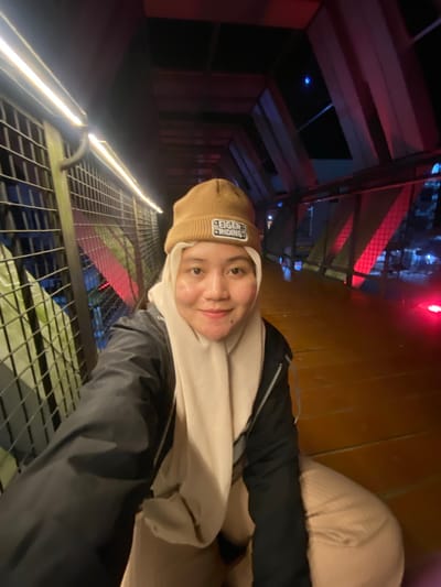 This is a selfie taken by a person wearing a tan beanie and a light-colored scarf or hoodie. The person has a gentle smile. They are wearing a black jacket over a light-colored garment. The background shows an indoor or covered walkway with a metal grate floor and railing. There are illuminated lines along the walkway and some reddish-pink lighting in the distance, suggesting an entertainment venue or a pathway with decorative lighting. The time of day appears to be evening or night due to the artificial lighting. The texture of the wooden wall on the right side of the image is visible. The text "EIGER RIDING" is visible on the beanie. The location context is Banjarbaru, Indonesia.