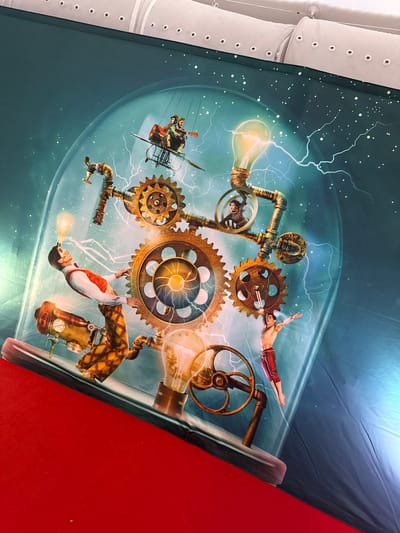 This image depicts a vibrant and fantastical scene with a steampunk aesthetic. The central focus is a collection of oversized gears and intricate pipework, suggesting an elaborate mechanical contraption. Several individuals are integrated into this machinery. One person, a man, is suspended in mid-air, appearing to be performing an acrobatic feat. Another group of people are seated in what looks like a rudimentary flying machine, also suspended amidst the gears. The overall scene is illuminated by a bright, glowing lightbulb, and streaks of lightning add a dramatic and energetic element. The backdrop is a swirling, gradient blue, reminiscent of a nebula or a stylized sky, dotted with small white specks that could represent stars or energy particles. The image appears to be a graphic or artwork, possibly a poster or a mural, given the smooth, untextured appearance. There is no visible text in the image. The mood is one of wonder, adventure, and ingenuity, characteristic of steampunk art. The scene is set within a transparent dome-like structure, further enhancing the fantastical and contained nature of the display.