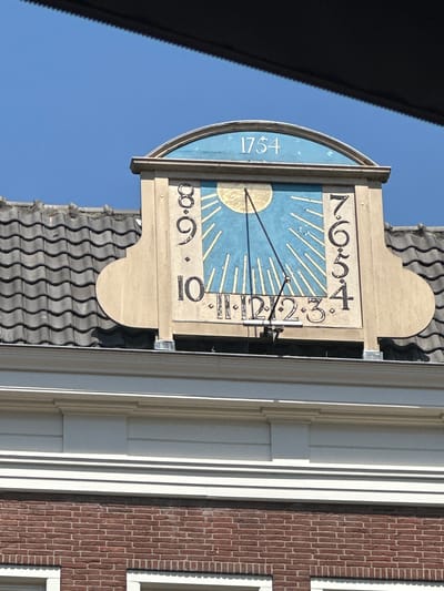 The image shows a sundial on the roof of a building, likely in Zwolle, Netherlands. The sundial is framed by beige, ornate woodwork and features a blue face with golden sun rays. The year "1754" is inscribed in white above the blue face. The hours are marked in black numerals, with "8, 9, 10" on the left side and "7, 6, 5, 4" on the right. The sundial also has Roman numerals "XI, XII, 12, 2, 3, 4" along the bottom. A thin, black gnomon casts a shadow across the dial. The sundial is situated above a grey, tiled roof and below a clear blue sky. The building below the roof is made of red brick and has white trim. The photo appears to be taken from a slightly elevated position, possibly from another building or a drone.