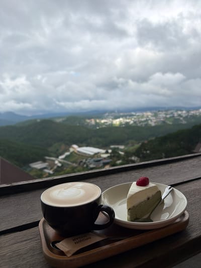 A slice of cake with a strawberry on top sits on a white plate next to a cup of latte art coffee, both placed on a wooden tray. The tray is on a dark wooden table. In the background, a hazy cityscape with rolling green hills under a cloudy sky is visible. The scene is likely from a cafe or restaurant in Xuân Thành, Vietnam, offering a scenic view.