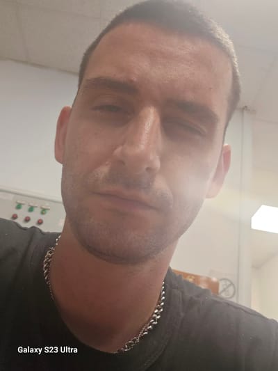 The image is a close-up, selfie-style photograph of a man's face, taken indoors. The man has short hair and is wearing a dark t-shirt and a silver chain necklace. He appears to be looking down with his eyes partially closed, and there is a noticeable glare on his forehead and cheek. The background is blurred, showing what appears to be an industrial or utility setting with some machinery or control panels on the left and a bright light fixture or window on the right. A watermark in the bottom left corner reads "Galaxy S23 Ultra". There is no discernible information in the image to suggest a specific location in Haskovo, Bulgaria.