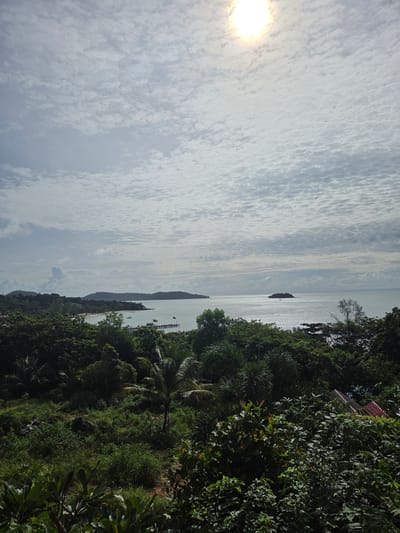The image depicts a panoramic view of Koh Toch Village, Cambodia, overlooking a tranquil bay under a partly cloudy sky. The sun, partially obscured by clouds, shines brightly in the upper right quadrant. The foreground is dominated by lush, dense green foliage, including palm trees and various tropical plants.
In the midground, a sandy beach curves along the coastline, dotted with a few scattered buildings and a pier extending into the calm, shimmering water. Several small boats are anchored in the bay. Further out, a small, tree-covered island stands in the distance. The sea extends to the horizon, where it meets the sky. The overall scene is one of natural beauty and coastal serenity.