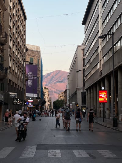 The image depicts a street scene in Palermo, Italy, during the late afternoon or early evening, as indicated by the warm, soft lighting on the distant mountains. The street is lined with tall, imposing buildings on both sides, characteristic of an urban environment. On the left, an older building with multiple windows and balconies is visible, while the right side features a modern building with a grid of windows and a columned facade.
The street itself is paved and appears to be a pedestrian thoroughfare or a street with limited vehicle traffic, as evidenced by the presence of a crosswalk in the foreground. Several people are walking along the street in various directions. In the immediate foreground, a man is riding a black scooter, passing by a pedestrian. Further down the street, groups of people are seen strolling, some with backpacks, suggesting they might be tourists or locals out for an evening walk.
Along the sides of the street, there are shops and businesses. A large purple banner with Italian text and a QR code hangs from one of the buildings, likely an advertisement. Another prominent advertisement, a bright red digital screen displaying text in yellow, is positioned on the right side of the street. The background is dominated by a view of mountains, illuminated by the setting sun, which provides a natural and scenic backdrop to the urban setting. The sky is clear and a pale blue, fading to a lighter hue near the horizon. Overhead, wires are strung across the street, a common sight in urban infrastructure.
