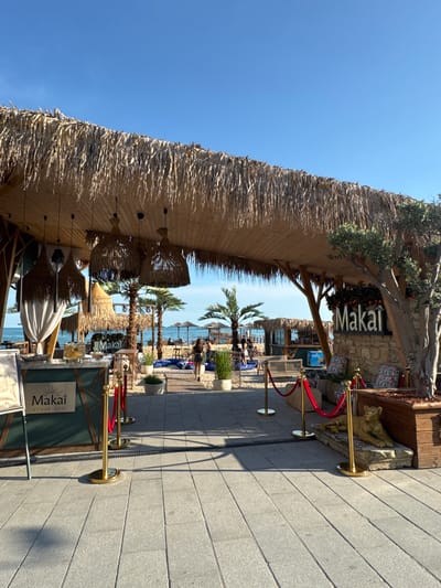 This image depicts the exterior of a beach bar named "Makai" in Balchik, Bulgaria. The establishment features a thatched roof, wooden beams, and straw-woven hanging lamps. In the foreground, a green bar with the "Makai" logo is visible, bordered by golden stanchions and red velvet ropes. Behind the bar, an olive tree stands next to a stone facade displaying the "Makai" sign. The background reveals a beach with palm trees, lounge chairs, and umbrellas, facing the Black Sea. Several patrons are seen on the beach and walking towards the bar. The sky is clear and blue, suggesting it is a sunny day.