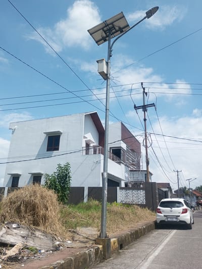 A street scene shows a solar-powered street light on the left, with a rectangular solar panel above and a boxy component below. A modern, white, two-story building stands behind the street light. To the right of the building, a utility pole is visible with numerous wires extending from it. A white car is parked on the side of the street. In the foreground, dry, dead grass and scattered trash clutter the area between the curb and the building. The sky is partly cloudy, showing blue patches interspersed with white clouds. The area does not appear to be in Zeyathiri District, Myanmar. A car license plate is visible, reading "F 1124 ZU."