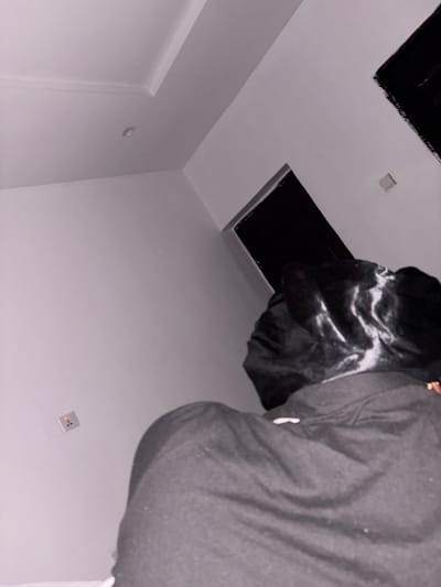 A person wearing a black satin hair bonnet and a gray shirt is looking up into a black, square-shaped hole in a white ceiling. The walls are also white, and there is a small square light fixture in the ceiling. The setting appears to be indoors. There is no information to identify this scene to Iddo Tudunwada, Nigeria.