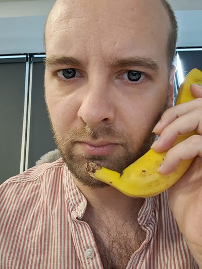 The image is a close-up, rotated selfie of a person holding a banana. The person has light skin, blue eyes, and a light brown beard. They are wearing a red and white striped shirt. The background appears to be indoors, with a gray surface and some horizontal elements that could be part of a desk or shelf. The lighting suggests it is daytime. The person's expression is neutral. No visible text is present in the image.