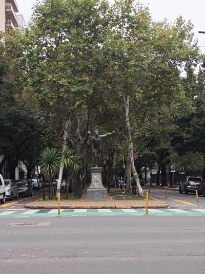 The image depicts a street scene in La Plata, Argentina, dominated by a statue of a nude male figure with arms outstretched, situated in the center of a traffic island. The statue stands atop a stone pedestal. Behind the statue are tall, leafy trees lining the median strip of the street. Two small palm trees flank the base of the statue. Several cars are parked on either side of the street. Yellow bollards mark a green-and-white crosswalk in the foreground. The sky is overcast. Buildings are visible in the background.