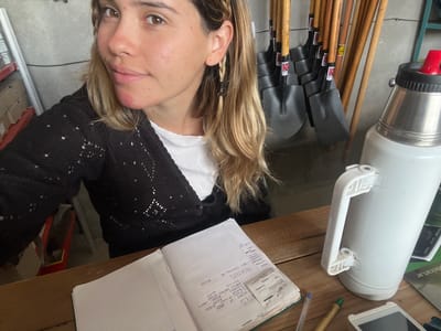 A selfie taken by a person with light skin and brown eyes, wearing a white shirt and a black crocheted cardigan. The person has blonde hair, partially braided, and is facing the camera. The background consists of a cluttered workshop or storage area.
Several shovels with wooden handles are leaning against the wall. Next to them is a large white thermos with a red spout. A notebook is open on a wooden surface, with handwritten notes and a receipt, likely recording expenses. Some of the handwritten notes are for "MARTES" (Tuesday) and "MIERCOLES" (Wednesday). Other objects on the wooden surface include pens and a small electronic device. Based on the text "Sumalao" and "oxiran.ar" printed on a receipt, the location is identified as Sumalao, Argentina.
