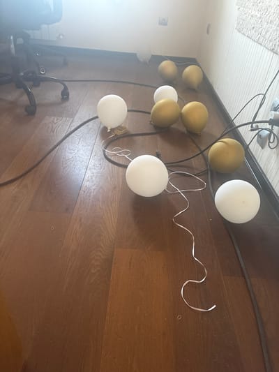 The image displays a cluster of deflated balloons and electrical cords on a wooden floor. The balloons are of two colors: white and gold. Some balloons are lying flat, while others appear partially inflated. The electrical cords are black and snake across the floor, connecting to what appear to be power outlets or fixtures on the wall in the background. A portion of an office chair with wheels is visible in the upper left corner. The wall in the background is light-colored and textured. The lighting in the scene suggests an indoor environment, possibly a room in a residential or office building. The location is identified as Orekhovka, Russia.