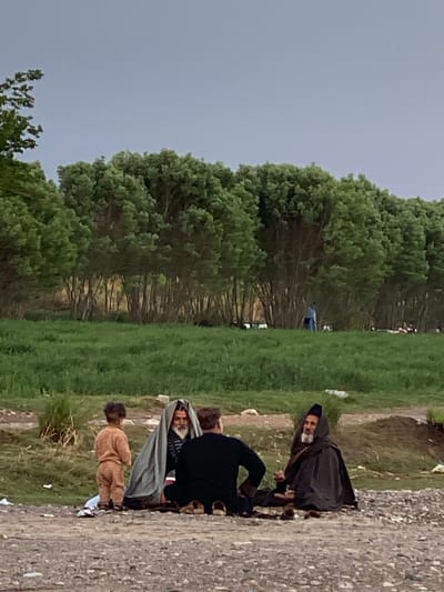 The image displays an outdoor scene in Arghandab, Afghanistan, under an overcast sky. In the immediate foreground, a gravel surface hosts four individuals. Three adult males are seated: one on the right wears a dark hooded cloak and has a white beard; the central male is partially covered by a light grey fabric over dark clothing, also exhibiting a white beard; and the male on the left is observed from behind, dressed in a dark long-sleeved top. A small child, wearing a light brown outfit, stands facing away from the viewer towards the seated adults. Scattered debris is visible on the gravel. The midground consists of a vibrant green grassy field, which extends to a dense line of tall, leafy green trees forming the background. Within the midground field, near the tree line, an individual in light blue apparel is visible standing. The seated adults appear to be engaged in a social gathering.