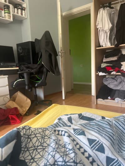 The image, taken in Pokrovnik, Bulgaria, captures an indoor room, likely a bedroom, with a glimpse into an adjacent space. The perspective is slightly tilted, showing the room from an elevated angle.
On the left side of the frame, an open wardrobe or shelving unit is visible. The upper shelves hold a mix of dark and colorful garments, while the lower shelf contains a stack of neatly folded white t-shirts or similar apparel.
Towards the center, a white door frame leads into what appears to be a hallway or another room, distinguished by a vibrant lime-green wall. A white door, slightly ajar, features a dark handle.
The right side of the image is dominated by a bed, partially covered with a bright yellow sheet. A patterned duvet, featuring intricate white and blue/grey geometric designs, including stripes, triangles, and abstract lines, is draped over the bed. Below the bed level, a black gaming chair with green accents is positioned next to a black PC tower and a monitor/screen. Adjacent to this setup, white wall-mounted shelves hold various bottles and containers, one of which clearly displays the text "CREATINE". On the floor, a brown paper bag with the partial text "GAMON" is visible, alongside some crumpled red fabric.
No people are present, and no specific activity is occurring. The lighting suggests an indoor setting during daylight hours.