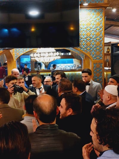 A group of people, mostly men, are gathered indoors, seemingly watching something. They are formally dressed, many in suits. The background features ornate, patterned decorations in blue and yellow, and a large, bright chandelier. A large dark screen is visible on the left. Several individuals are looking downwards, focused on an unseen object or event in front of them. A photographer holds a camera towards the scene.