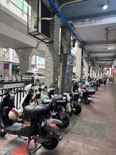 The image depicts a street scene in Yuexiu District, China, characterized by a row of parked electric scooters and motorcycles lining a brick-paved walkway. An elevated structure, possibly part of a bridge or overpass, casts a shadow over the scene. The architecture includes pillars with a brick facade and exposed concrete. Several air conditioning units are visible mounted on the exterior walls and under the elevated structure. A light-colored SUV is parked in the background, behind a white fence. In the distance, a woman in a pink shirt walks along the walkway, and other pedestrians are present. The overall environment suggests a public urban space with a focus on transportation and commercial activity, as indicated by storefronts visible in the background.