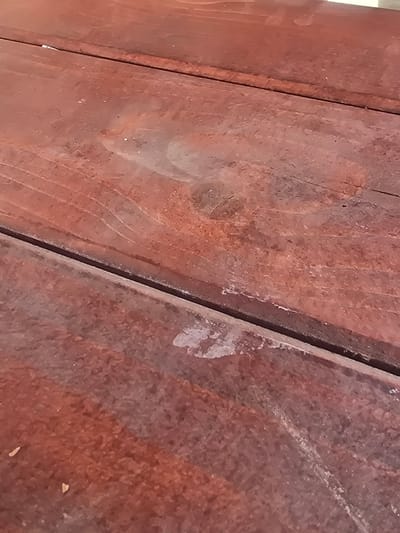 The image captures a close-up, downward-angled view of a horizontal, reddish-brown wooden surface located in Nyíregyháza, Hungary. The surface consists of at least three distinct wooden planks, separated by dark, narrow linear gaps. The wood displays significant wear, characterized by numerous irregular light-colored scuff marks, areas of eroded finish, and scattered white particulate matter consistent with dust or debris. Fine linear abrasions are visible across various sections. No individuals, specific objects beyond the wooden surface, or discernible actions are present within the frame.